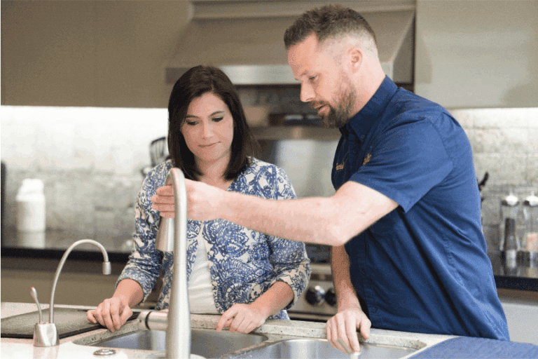 A man shows a woman how to use a kitchen faucet in a modern kitchen setting.